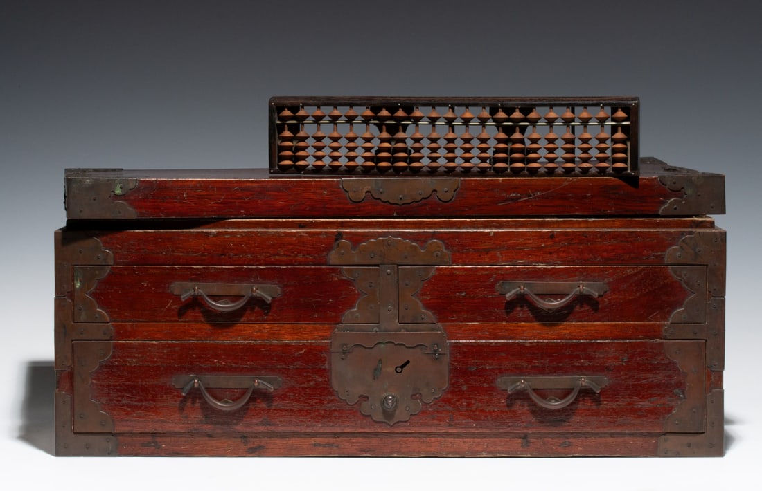 A MEIJI PERIOD JAPANESE MERCHANT'S CASE WITH ABACUS: The paulownia wood case of drawers is finished in an old red stain and finished with darkly patinated bronze or copper mounts featuring an upper compartment with lift top and two half-drawers over