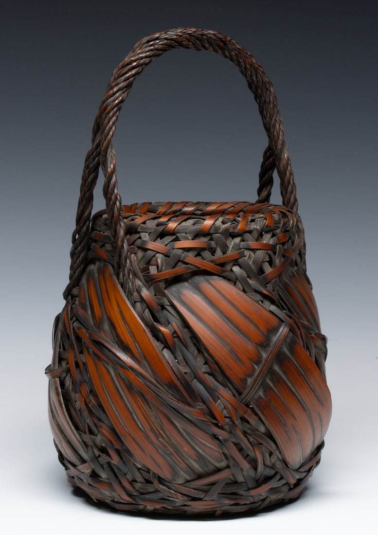 A JAPANESE BROAD SPLINT IKEBANA BASKET, ADAIR ESTATE: The bold and expressive example is dominated by wide, curved bamboo splints interlocked within a complex random-weave joining a thick, tightly twisted bamboo braid handle. Measures 14.5 x 8.25 x 8.25 