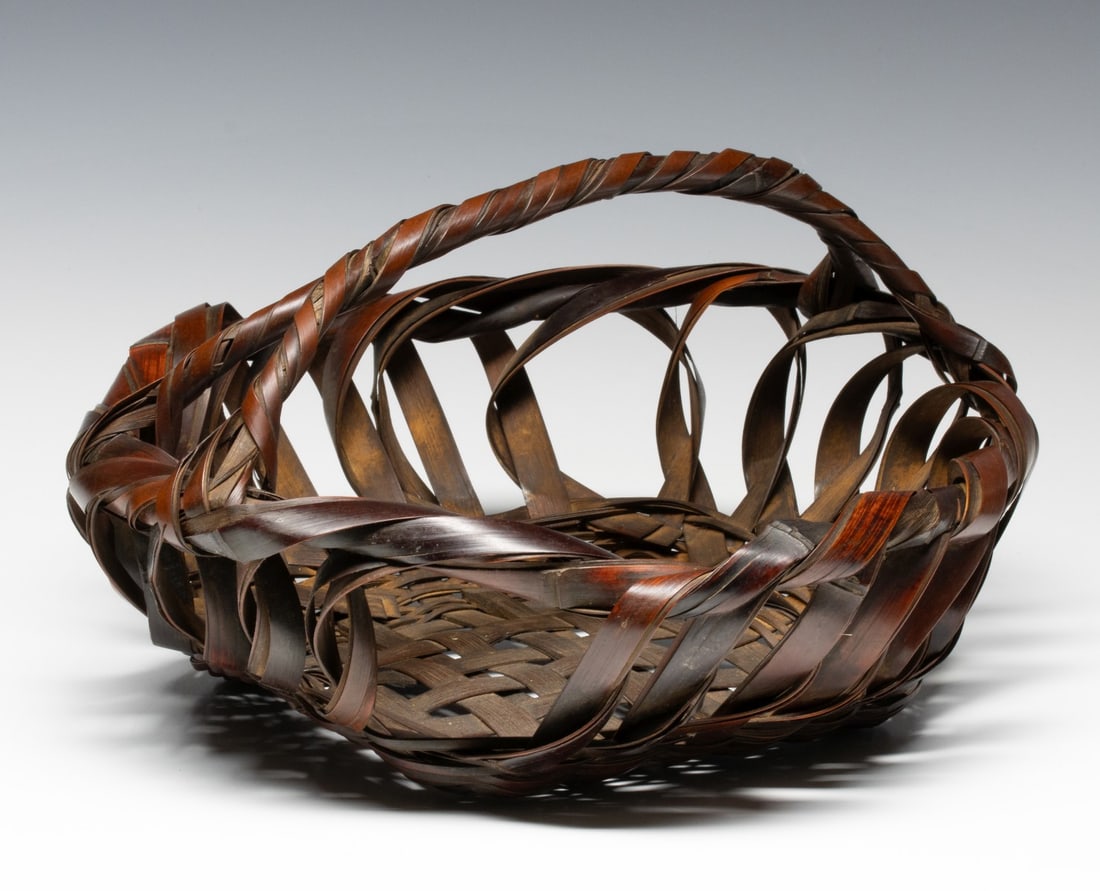 A BOWL FORM JAPANESE BAMBOO FLOWER BASKET, ADAIR ESTATE: The low, circular form basket with handle is woven to create a dynamic and disorderly character in rustic overtones. Measures 7.75 x 14.25 x 14 inches. Provenance: The estate of John Adair Jr. (1942-2