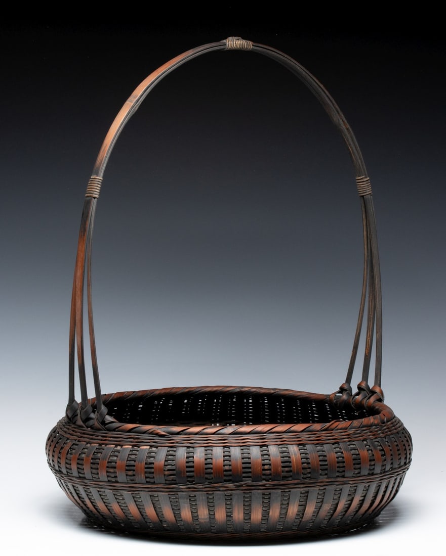 A JAPANESE BAMBOO BASKET FOR IKEBANA, ADAIR ESTATE: The wide, shallow with rope-twist rim has vertical-stay plaiting technique with wide, flat bamboo slats held in place by horizontal bands. The high handle is composed of three individual bamboo slats 
