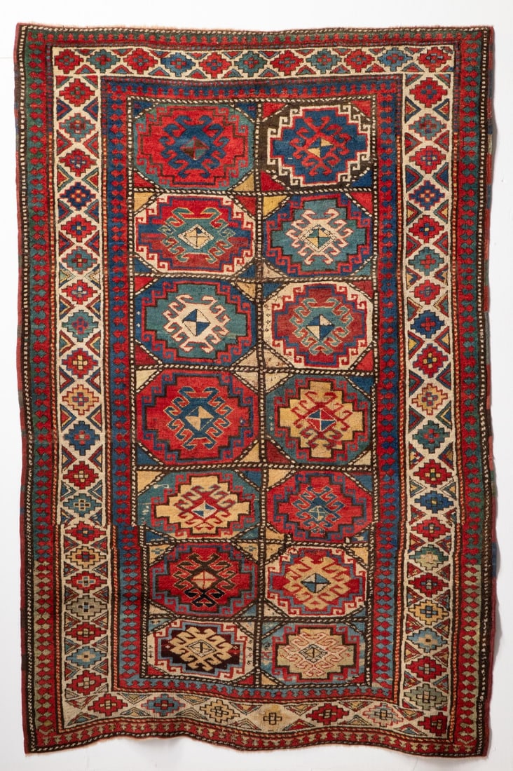 A 19TH CENT MOGHAN KAZAK RUG, WEST CAUCASUS, AS FOUND: The polychrome field with two columns of seven rectangular reserves, each enclosing a 'Memling' hooked and stepped medallion within an ivory border enclosing continuous stepped polychrome medallions. 