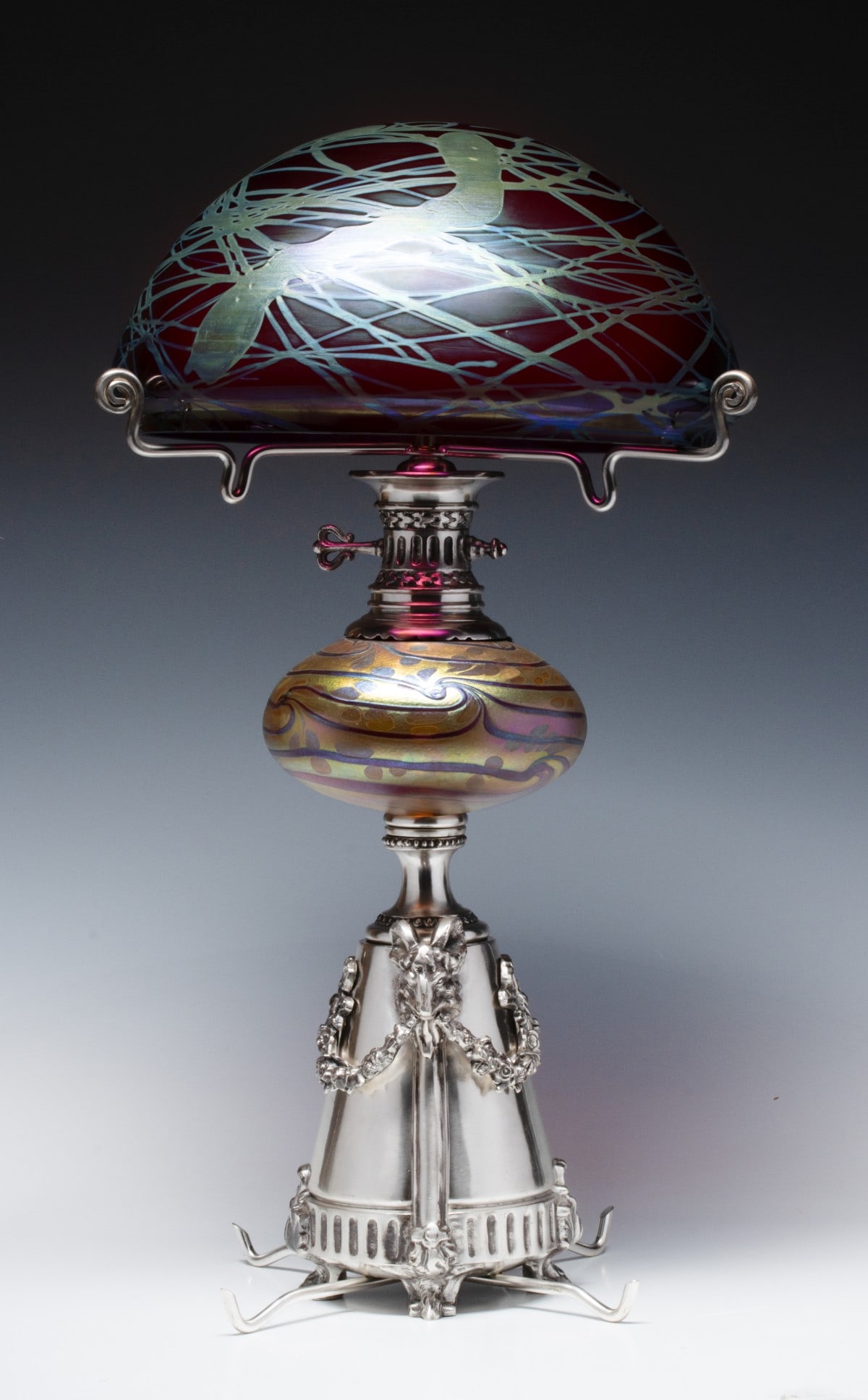 AN AUSTRIAN ART GLASS AND SILVER PLATE TABLE LAMP: The silvered base with ram's heads and garlands fitted with an oil spot and pulled thread art glass font attributable to Loetz, and a similar mushroom form Art Nouveau iridescent shade. Measures 23.5