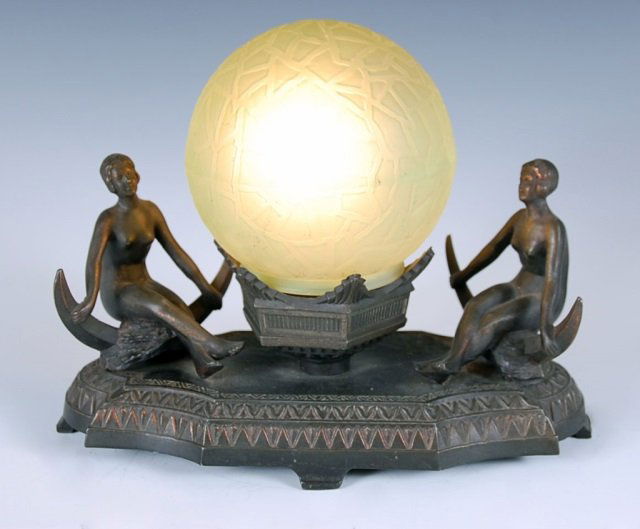 ART DECO NOVELTY LIGHT WITH TWO NUDES: Spelter base with figural nudes sitting on crescent moon at either side of green frosted shade. Measures 8 inches high by 11 inches.