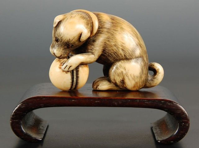 CARVED ASIAN IVORY OKIMONO OF PUPPY WITH BALL: A nice quality signed carving with inset ebony eyes. Measures 1.5 x 2 inches excluding stand. NO INTERNATIONAL SHIPPING OF IVORY.