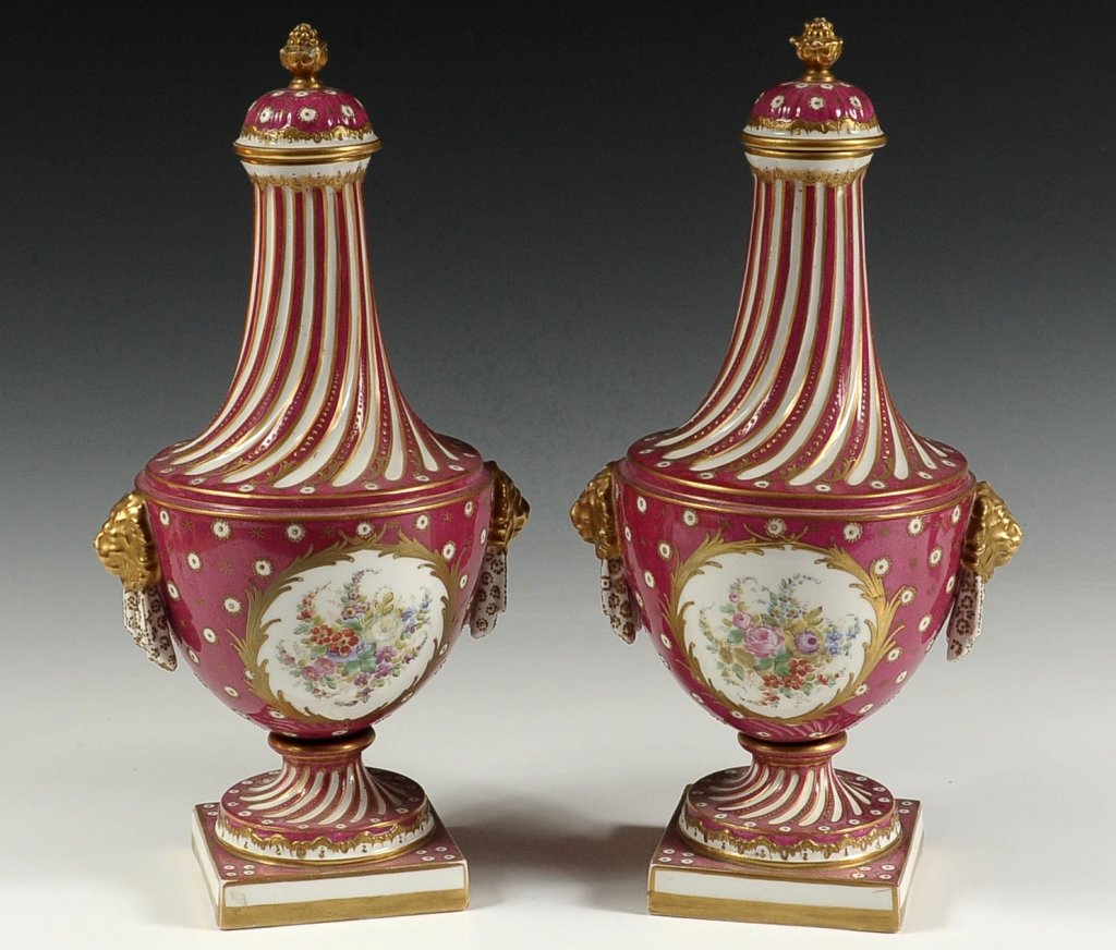 PAIR OF SEVRES FRENCH PORCELAIN BOLTED AND COVERED URNS: Pair of bolted urns with hanging flower basket decoration fully painted on one side and floral spray on the opposite side. All accented with raised gold enamels. A Sevres mark in blue over glaze on th