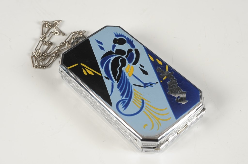 E.A.M. ENAMELED VANITY CASE WITH A BIRD DESIGN: Enameled bird on a silvertone vanity case with a chain handle, framed mirror in the lid, hinged comb in a clip, base holds a rouge well with pan and puff and a powder well with a rotary sifter, marked