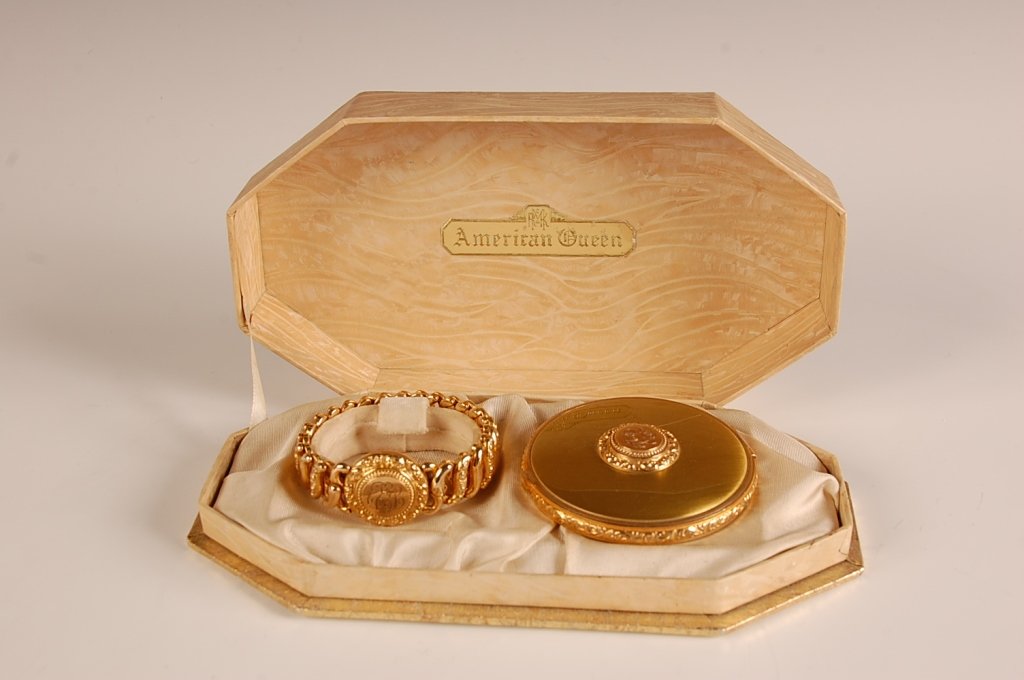 COMPACT WITH MATCHING BRACELET & PRESENTATION BOX: American Queen' compact with monogram decoration on top, hinged metal mirror in the lid over a rouge well with pan and puff, hinged powder well in the base with puff, unmarked. Matching goldtone brace