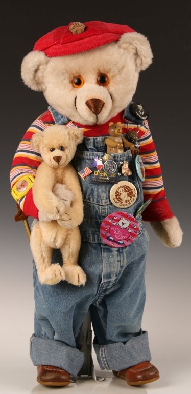 ARTISAN TEDDY BEAR BY BALLARD BAINES: Dressed in Osh Kosh overalls with cap, buttons and holding another teddy bear, measures 25 inches.