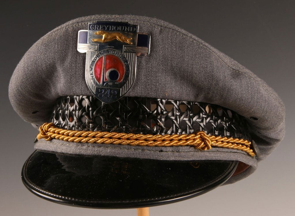 1939 GREYHOUND BUS WORLD'S FAIR VISOR CAP AND BADGE: Fine grey wool visor cap with caned side, gold braided chincord, with side buttons. Black patent leather visor. Fine die struck enamel painted badge with applied gilt greyhound, depicts the 'World of