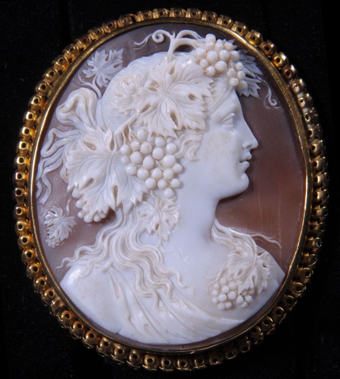 ANTIQUE CARVED SHELL CAMEO BROOCH IN 1OKT GOLD: A Bacchanalian theme Classical maiden with grape adornments. Carved shell in a 10kt gold setting, measures 2 x 1.75 inches.