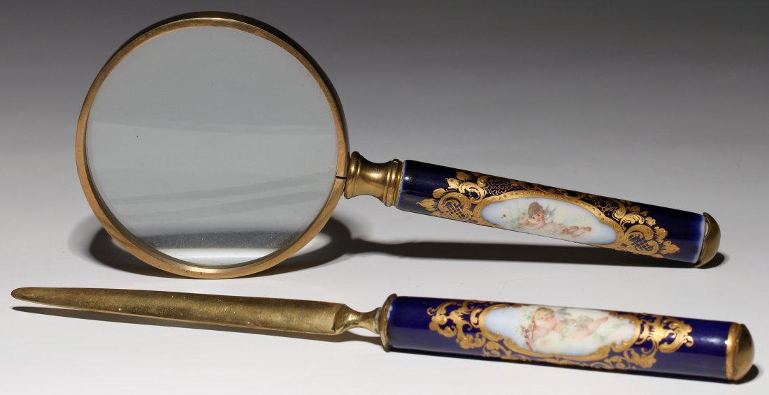 A SEVRES STYLE TWO PIECE DESK SET CIRCA 1900: Comprised of magnifying glass and letter opener, each of brass fitted with a cobalt blue glazed porcelain handle hand decorated with putti in a reserve surrounded by raised gold enamels. Measures 9.75