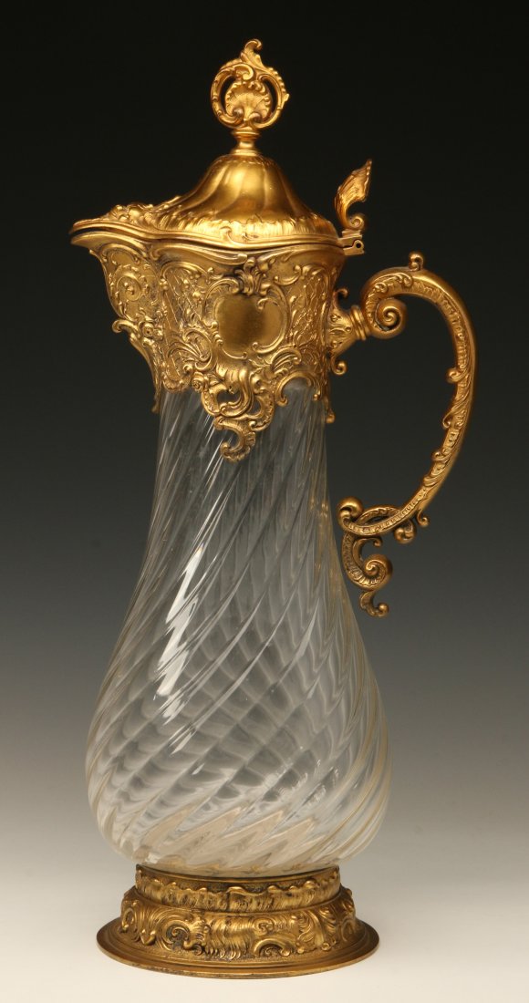 A CIRCA 1900 CRYSTAL FLAGON WITH GILDED METAL MOUNTS: The bulbous swirled glass body mounted with rococo style gilded metal foot and spout with handle and hinged cover. Measures 15.75 x 7 inches.