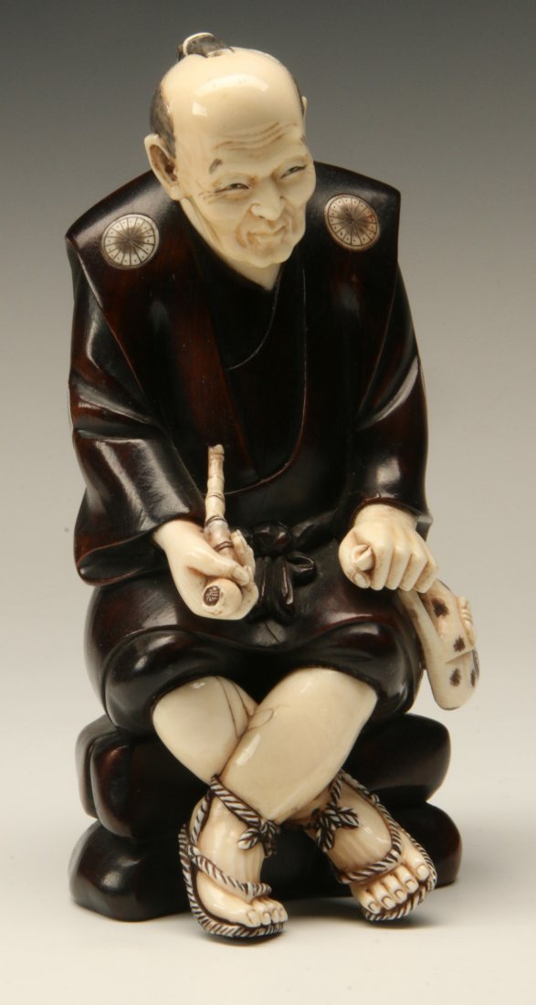 A MEIJI PERIOD JAPANESE IVORY AND HARDWOOD CARVING: A Japanese ivory and hardwood carving sensitively and meticulously carved depicting a figure of a seated man smoking a pipe and clutching a tobacco pouch; marked on base "Tani Azuma." Measures 6.5 hig