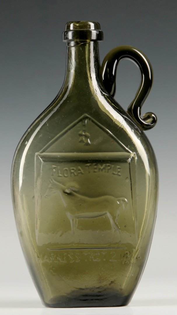 A RARE 19TH C. FLASK WITH RACEHORSE FLORA TEMPLE: A scarce blown glass flask fronted by an image of side view of a horse though stable doorway with tassels hanging above. Embossed lettering of 'Flora Temple Harness Trot 2.19 1/4', medium to dark gree