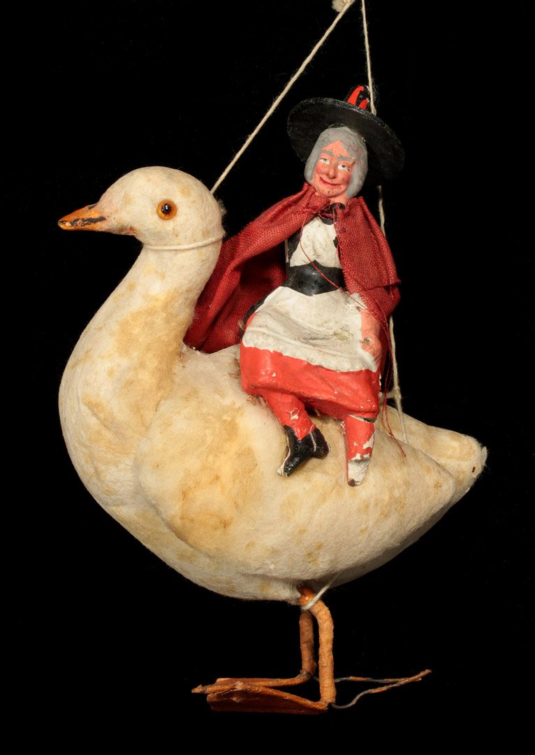 A CIRCA 1900 'MOTHER GOOSE' COTTON WOOL ORNAMENT: A cotton wool batting goose with glass eyes carrying a chalkware Mother Goose character with hand-painted details and a burlap cape, measures 5.5 x 4 inches.