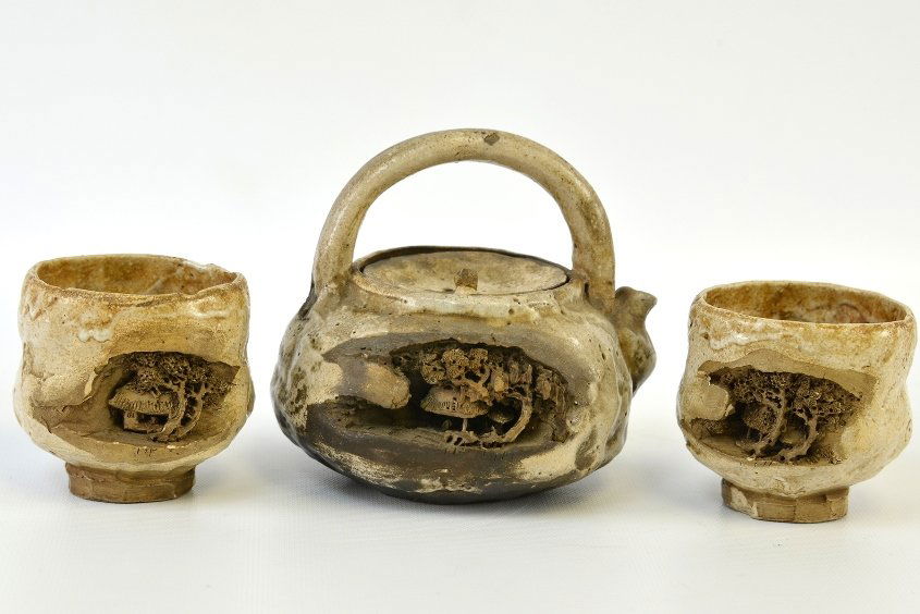 A Japanese Studio Ceramic Tea Set: A Japanese Studio Ceramic Tea Set, including a teapot and pair of cups, each with a recessed panel carved in high relief with a pavilion and tree scene, teapot appx. 4 1/2 in. H.