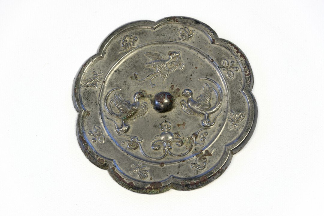 A Bronze "Birds and Flowers" Octalobed Mirror, Tang: Tang dynasty (618-907). Cast in relief to depict a bird in flight with a ribbon in its mouth above two mirrored perched phoenixes around a central pierced knob, the raised border cast with florets, in