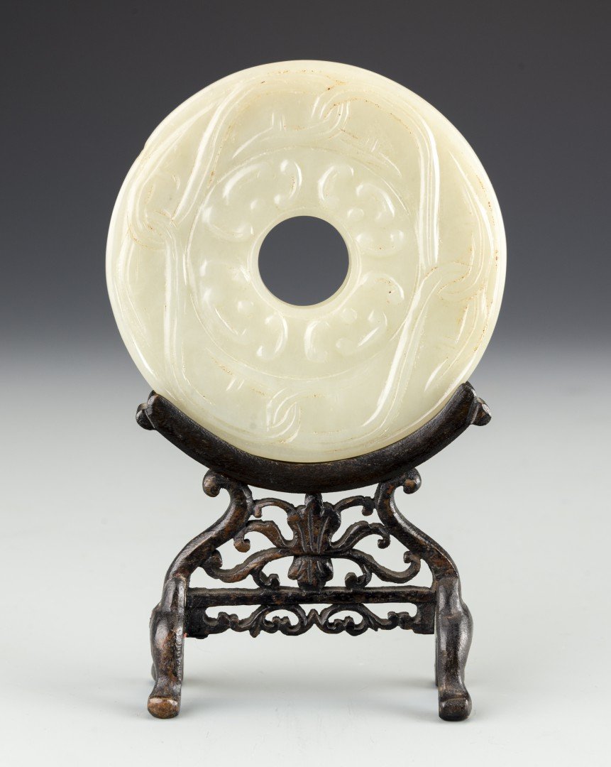 A Chinese White Jade Bi Disk in Wood Stand: 17th century, late Ming/early Qing dynasty. The white jade bi disk carved on both sides with interlocking tendrils and recumbent silk worm patterns. In a wooden stand with reticulated apron. Approx. 1