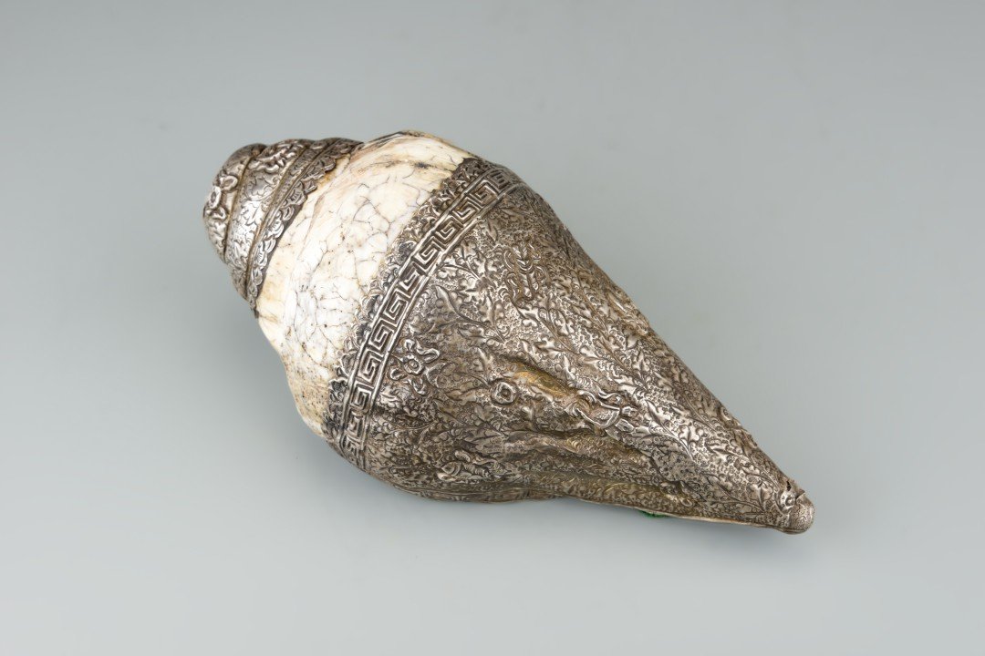 A Tibetan Metal Conch Shell Horn: A trumpet or bugle carved from a white conch shell with Tibetan silver overlay stamped in bas relief to depict floral meander and fish motifs. Conch shells are one of the Eight Buddhist Emblems and us