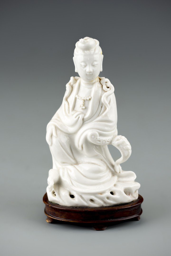 A Chinese Dehua Blanc de Chine Guanyin: The white glazed porcelain Guanyin is seated wearing jewels and robes. Fixed to a wooden base with "China" carved to the bottom. Approx. 6 by 3 1/8 in., 15.3 by 8 cm.