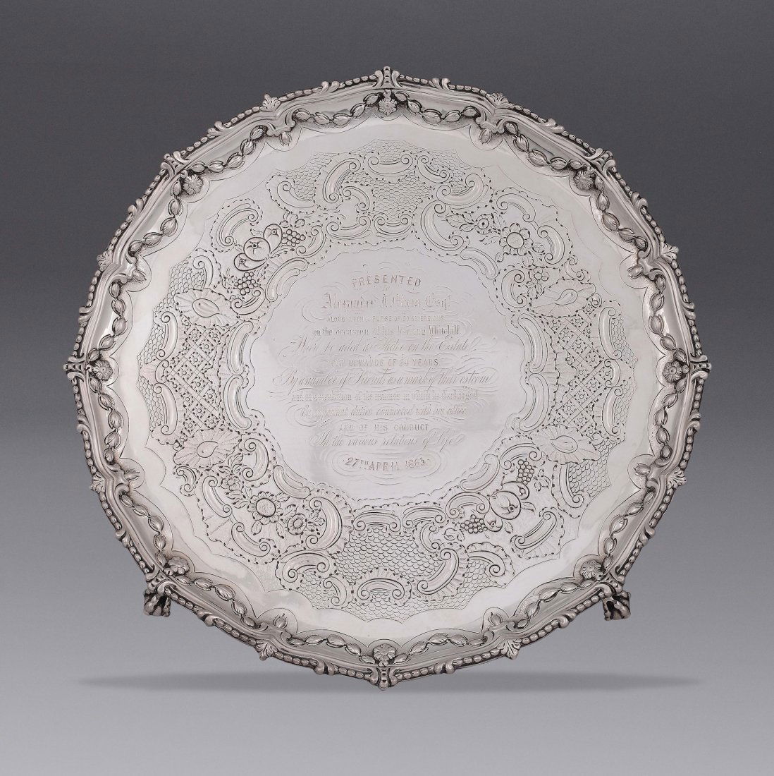 A silver Salver tray, London, 1771, maker John Carter: Salver circolare con bordo sagomato e decorato a perlinature e festoni fitoformi, cavetto riccamente cesellato a volute e riserve con al centro iscrizione incisa, piedini a motivo ferino gr. 1200, dia
