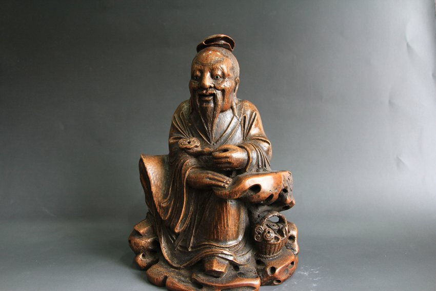 BAMBOO ELDERLY: Bamboo carved old man statue showing that he is sitting down along with a basket next to him. He is holding a Ruyi with his left hand. H: 19 cm, W: 14.5 cm