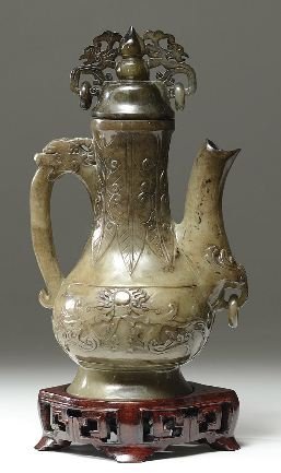 A CHINESE CARVED JADE TEA EWER: This well carved jade tea ewer rests firmly on a wooden stand. The kettle is carved with mythical images and has jade rings looped around the handle and stout. H: 9 5/8 in (24.5 cm), W: 4 1/2 in (11.5