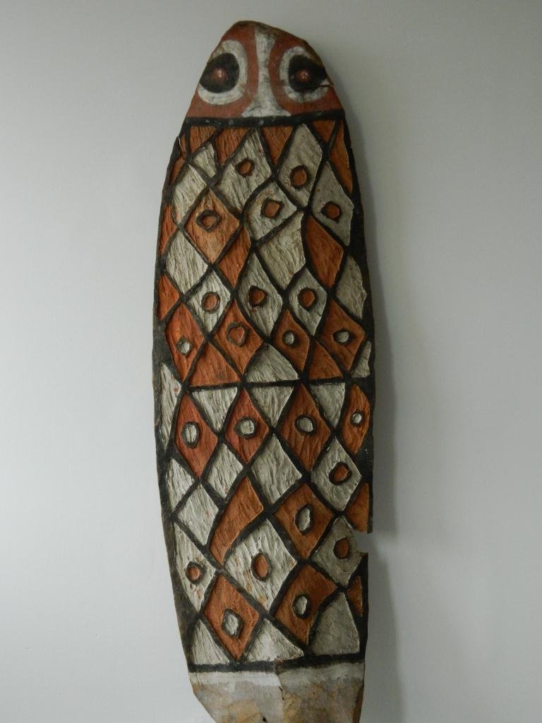 Papua New Guinea War Shield: #154 War Shield 62" muted red, black, and white paint is fading due to age, with a diamond design and a rudimentary face at top. It has a crack on top right side and a piece missing from lower right.