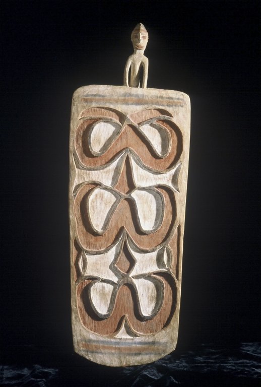 Papua New Guinea Asmat Shield: #182 39.75" high shield with red design on white background with a black horizontal stripe outlined in red at top and bottom of shield with a male figure carved on top,fading some.