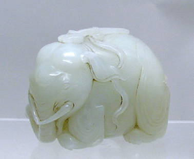 Chinese Carved White Jade Elephant: Finely carved elephant recumbent on four feet, head turned, and surmounted by a scepter with flowing ribbon. The jade a milky-white tone with opaque areas. 2 7/8"h x 3 ½"w.