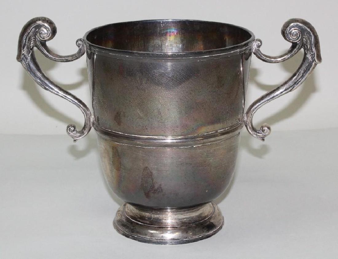 George II Twin Handled Loving Cup: C. 1710. Makers mark is illegible, bears Dublin purity mark and dates to 1710/11. Double handles are bifurcated C scrolls beginning & ending in swirls, the cup sits on a pedestal base. 6.75" H x 11"