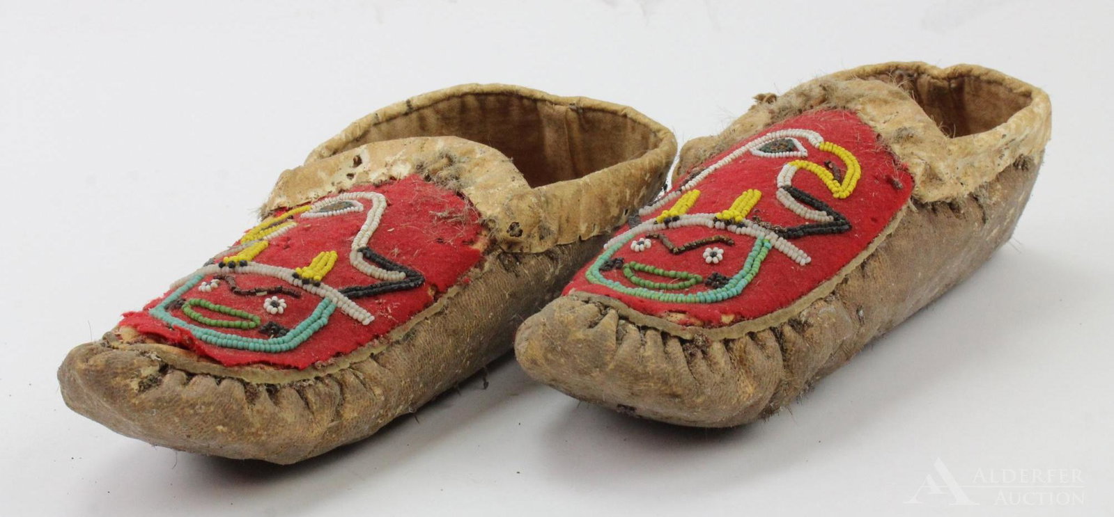 Pair Beaded Leather Moccasins: Cuffed hide moccasins with puckered toe. Red wool fabric on front with beaded eagle and face design.