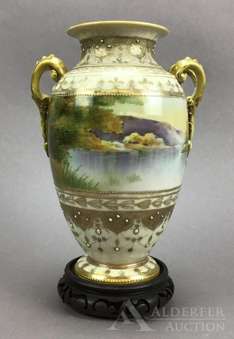 Morimura Nippon Vase: Morimura Brothers Nippon hand-painted double handled scenic urn/vase with water landscape scene and gold beading, measuring 10”h. With wood carved stand.