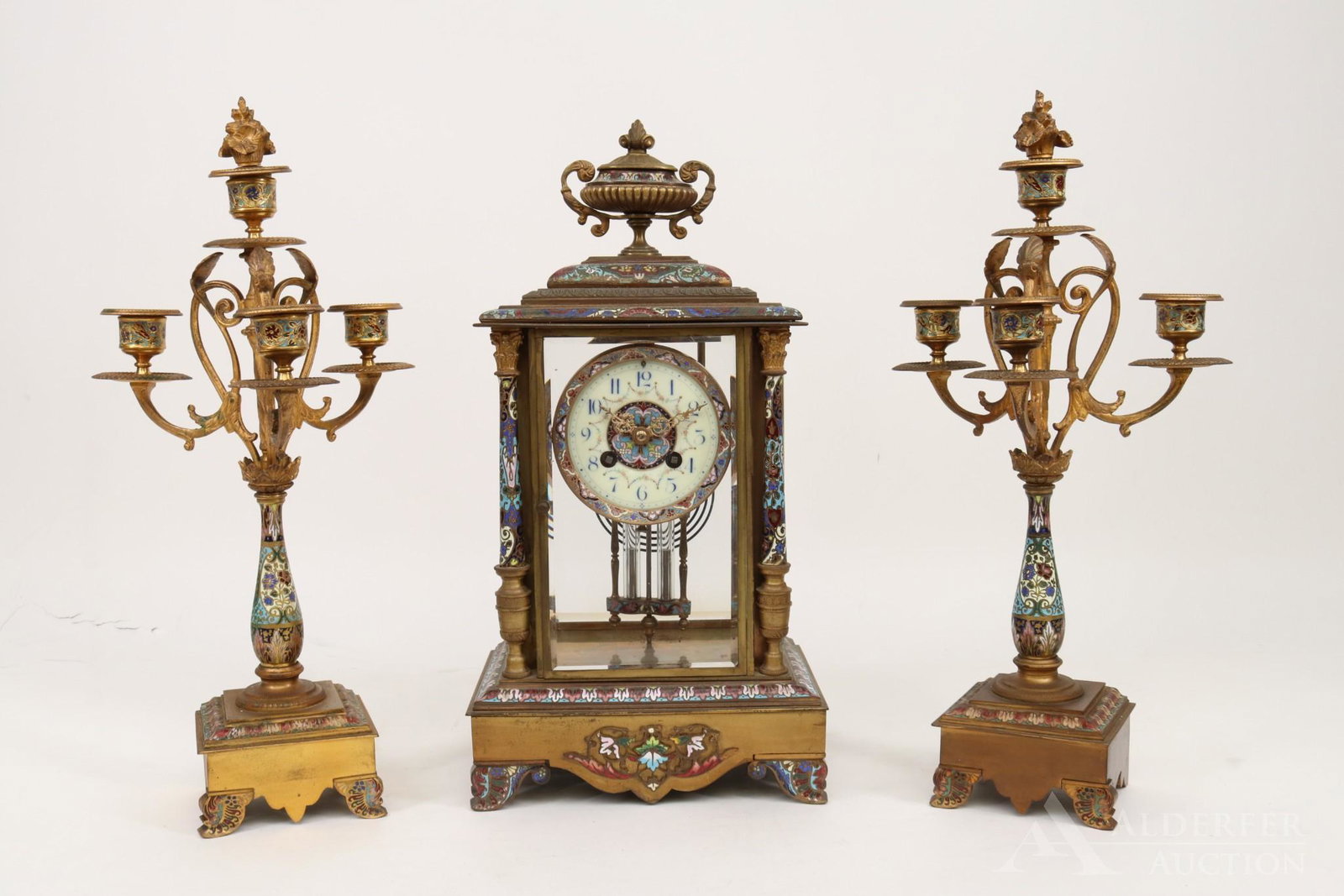 French Champleve Mantel Clock and Garnitures: Bronze champleve decorated mantel clock with urn finial and Corinthian columns. White enamel hand decorated face with blue Roman numerals and champleve decorated mercurial pendulum. Works marked Medi