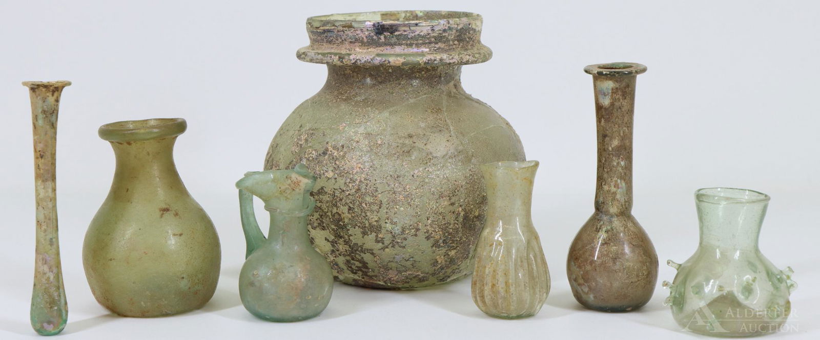 Roman Glass: Unguentaria / lacrymaria including a reeded example and an example with nodules, a small jug with folded lip and applied handle and a jar, total of (7) pieces of aqua and clear glass. Some show irid
