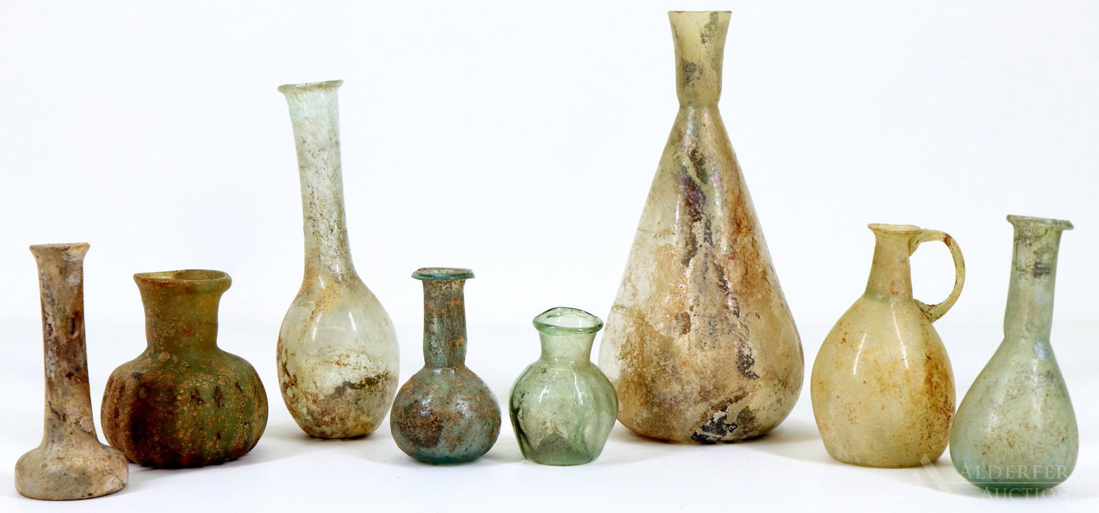 Roman Glass: Unguentaria / lacrymaria including an example with reeded body, and a jug with applied handle, total of (8) pieces of aqua and clear glass. Some show iridescence and other mineral deposits. Reconstr