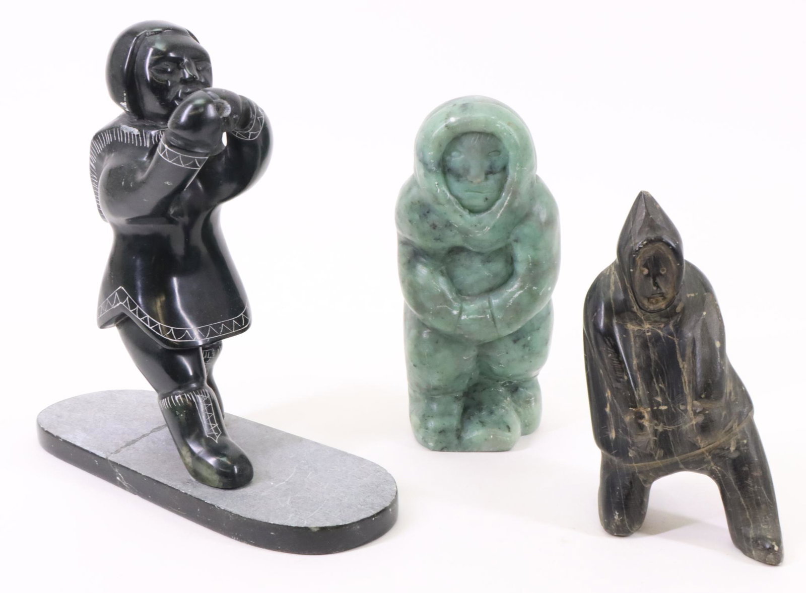 Inuit Carvings of Eskimos: Inuit carving of a standing Eskimo holding a fish, signed. Standing green Eskimo with a baby on their back, signed "W. Sharlow". Kneeling Eskimo. Largest measures 8”h x 8”w