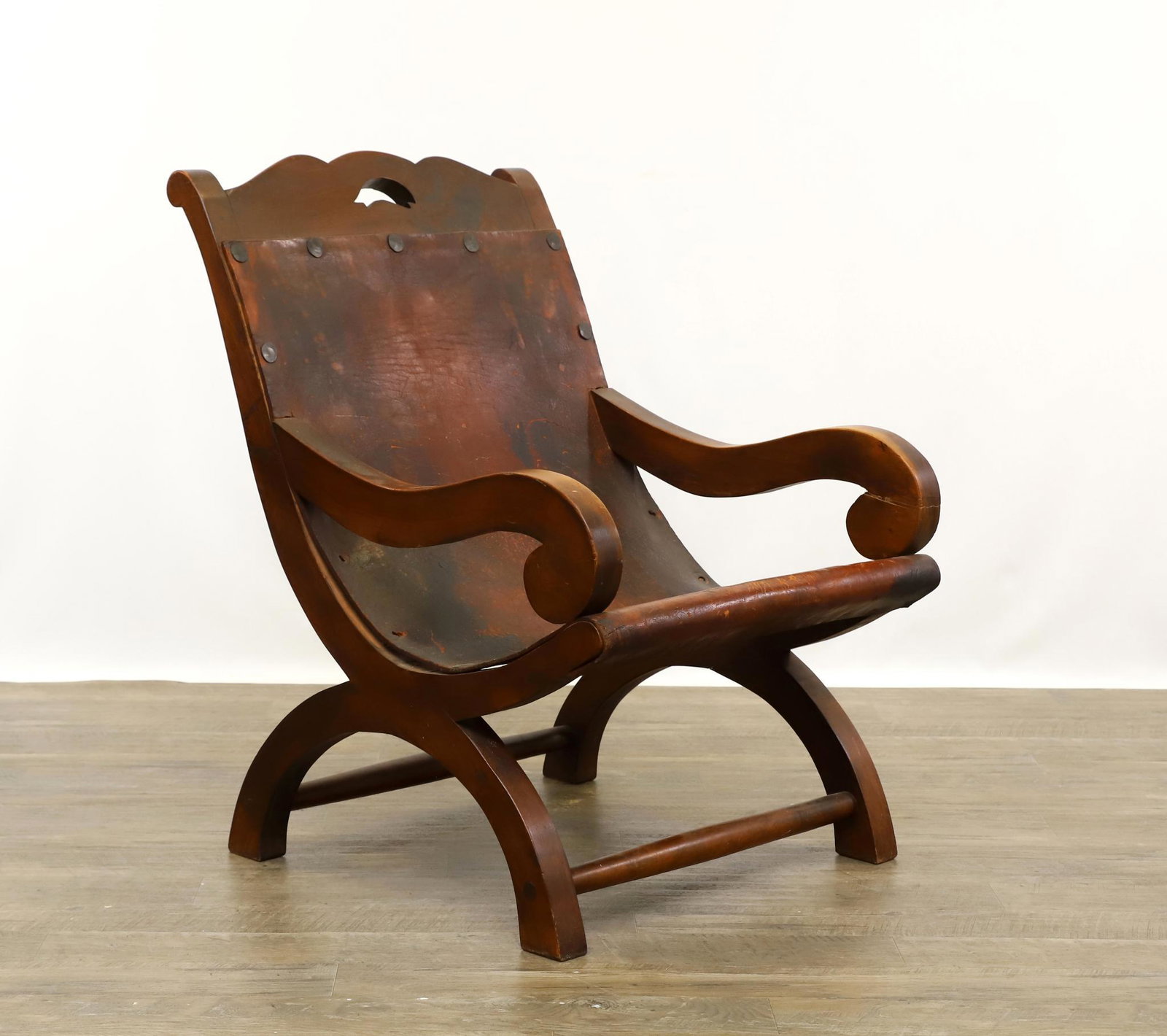 Campeche Chair In the Manner of William Spratling: Sabino x-frame chair, with scrolling arms, scalloped crest rail with pierced fish decoration, and leather sling seat held in place with hand wrought tacks, unmarked. Surface wear, repair to one arm, a