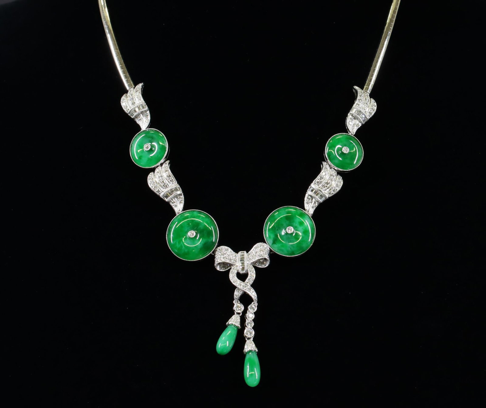 18KW Gold Jadeite and Diamond Necklace: Omega necklace in 18kw gold features a pattern of dark green jadeite disks and wings of diamonds culminating in the center with a bow of diamonds and ribbons with dangling tassels of diamonds and jade