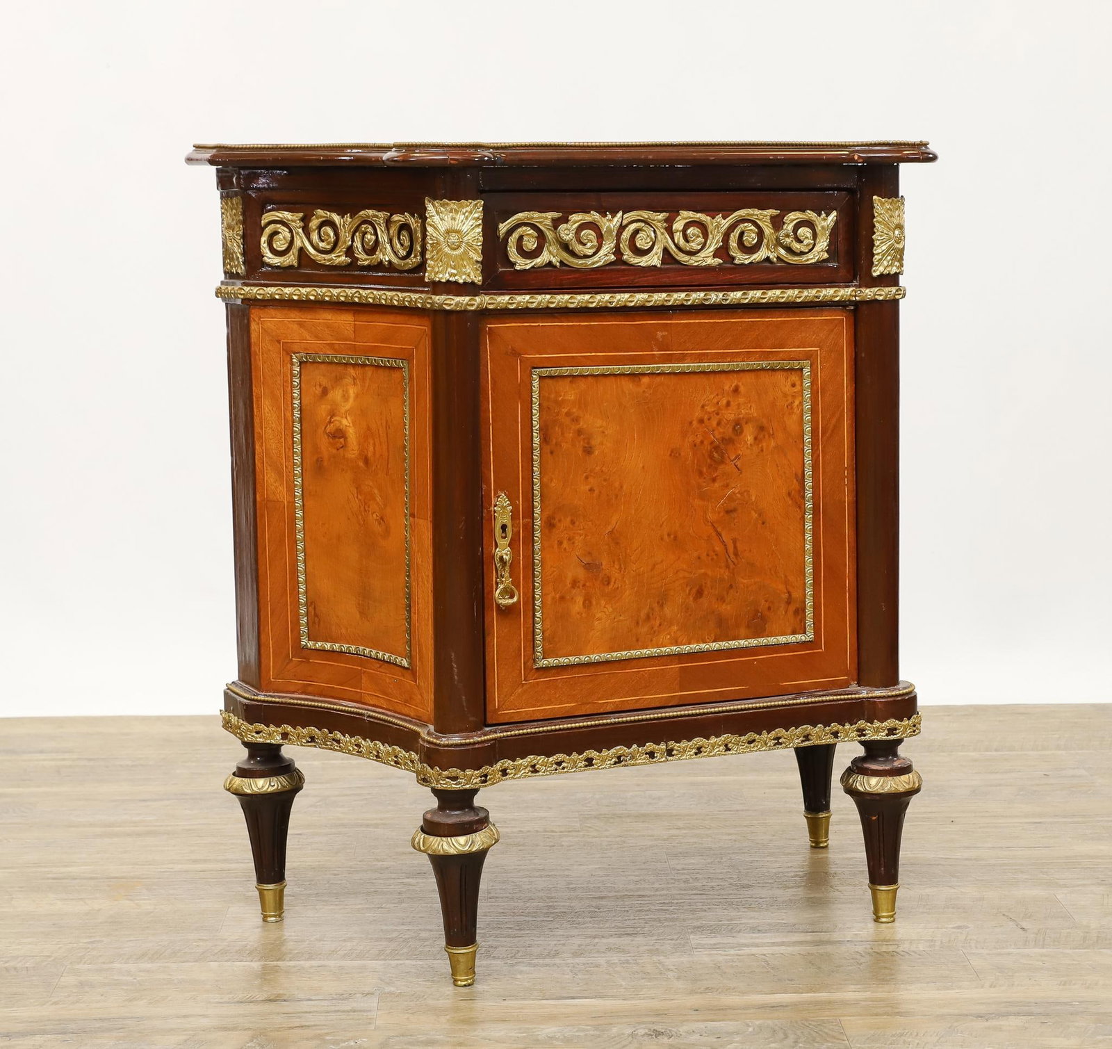French Ormoulu Mounted Side Cabinet: Satinwood, burl, and mahogany veneers having a concave top with cylindrical corners, one drawer over one door on turned toupe feet. No key is present with the door currently locked. 33"h x 30"w x 16"d