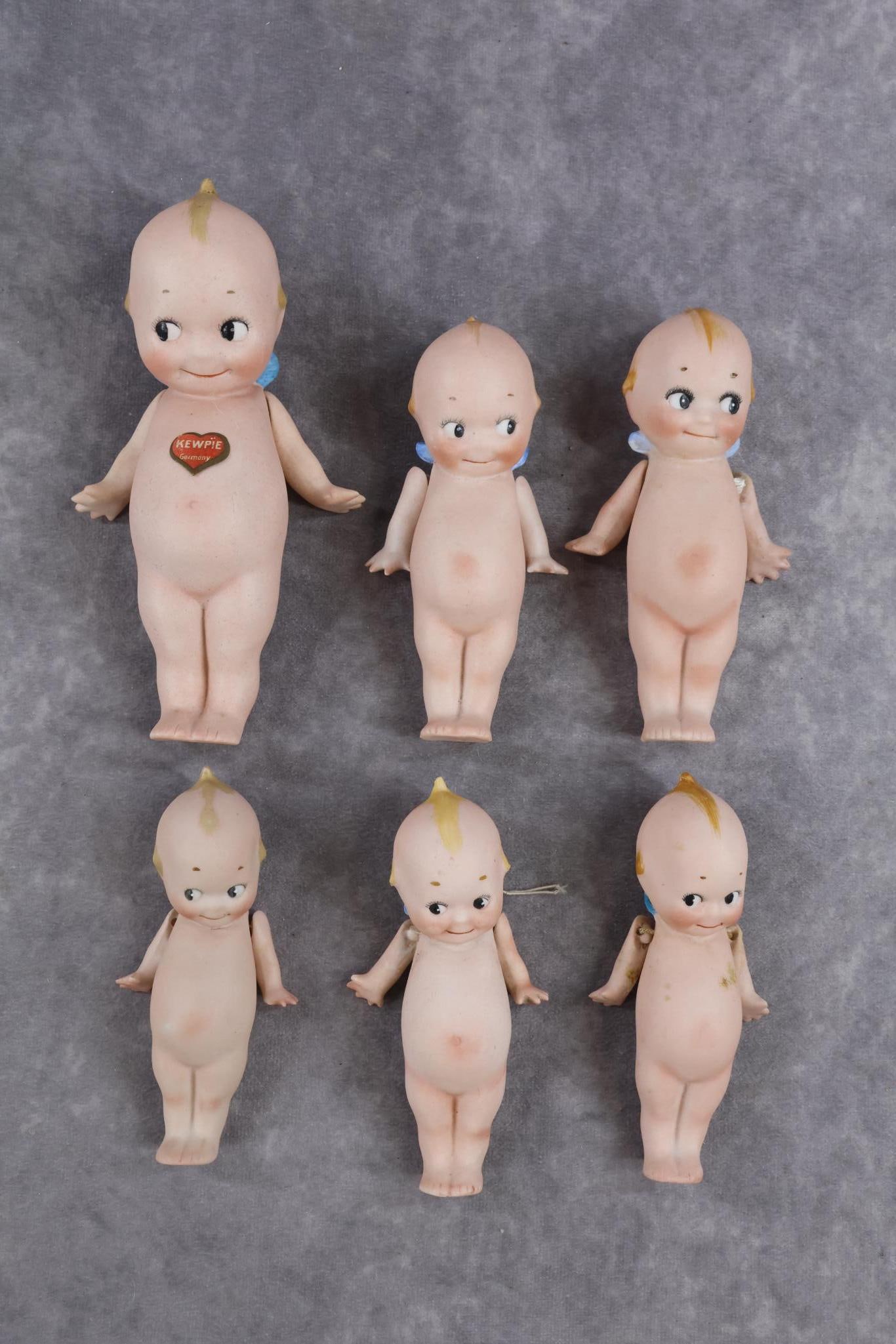 GERMAN ALL BISQUE DOLL(S).: LOT of (6) standing KEWPIE dolls. All have strung arms. (2) 4 1/2" Unmarked on feet. 4 3/4", (2) 5" Are all marked O'Neill on feet. (1) 5" Has a red "Germany' on back. The 6 1/2" Has the paper heart l