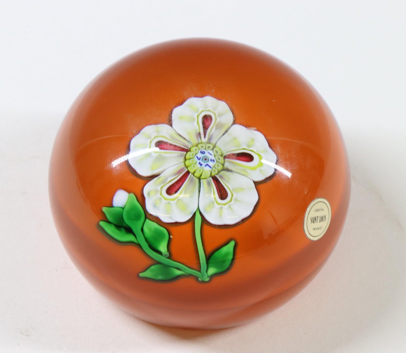 Saint Louis Crystal Paperweight: French lampwork paperweight decorated with a flower on an orange background. The cane at the center of the flower is marked SL 1973. Also retains a paper label. 2"h x 2.75"w