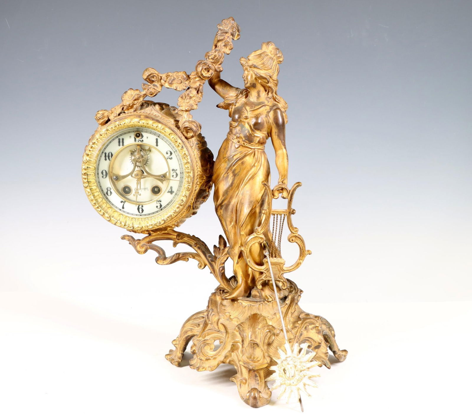 Gilbert Figural Clock: Art Nouveau gilt bronze and spelter sculptural clock, "Beatrice", a young woman with a harp and garland on a foliate base which also holds the case, featuring an open escapement and porcelain Arabic n