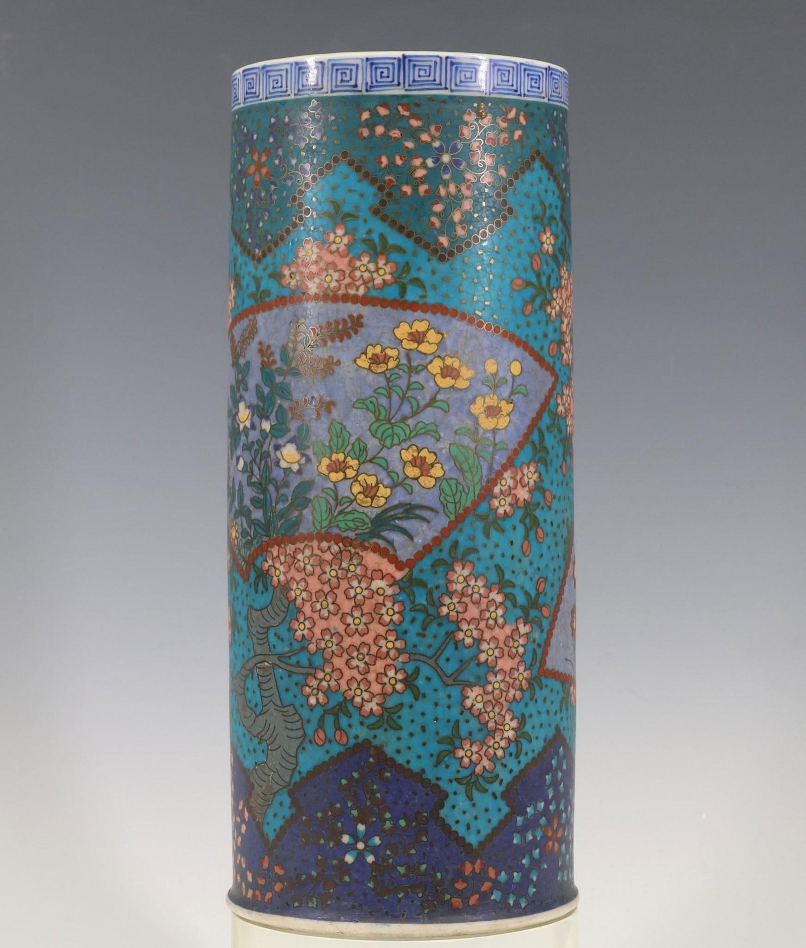 Japanese Totai Shippo Vase: Meiji era cloisonne on porcelain cylindrical vase decorated with panels depicting flowers and butterflies, with an underglaze cobalt meander top border. Hairlines, a chip to the foot, and other wear. 