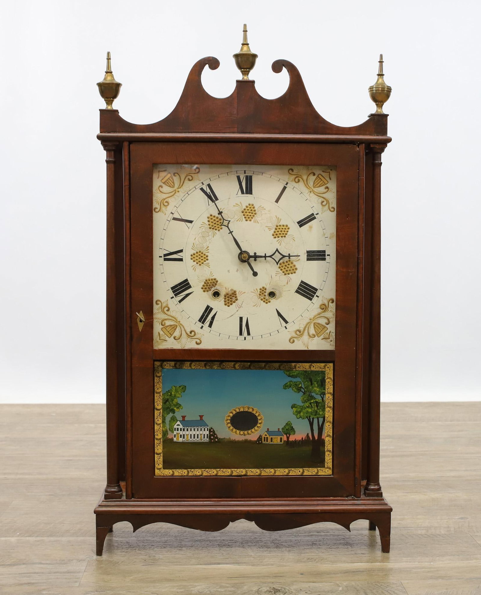 Elli Terry Pillar and Scroll Clock: Mahogany case, broken arch pediment with brass finials, turned columns, Hand painted and applied gilt enameled dial, reverse painted plantation panel, shaped apron on shaped feet. Weight and pendulum 