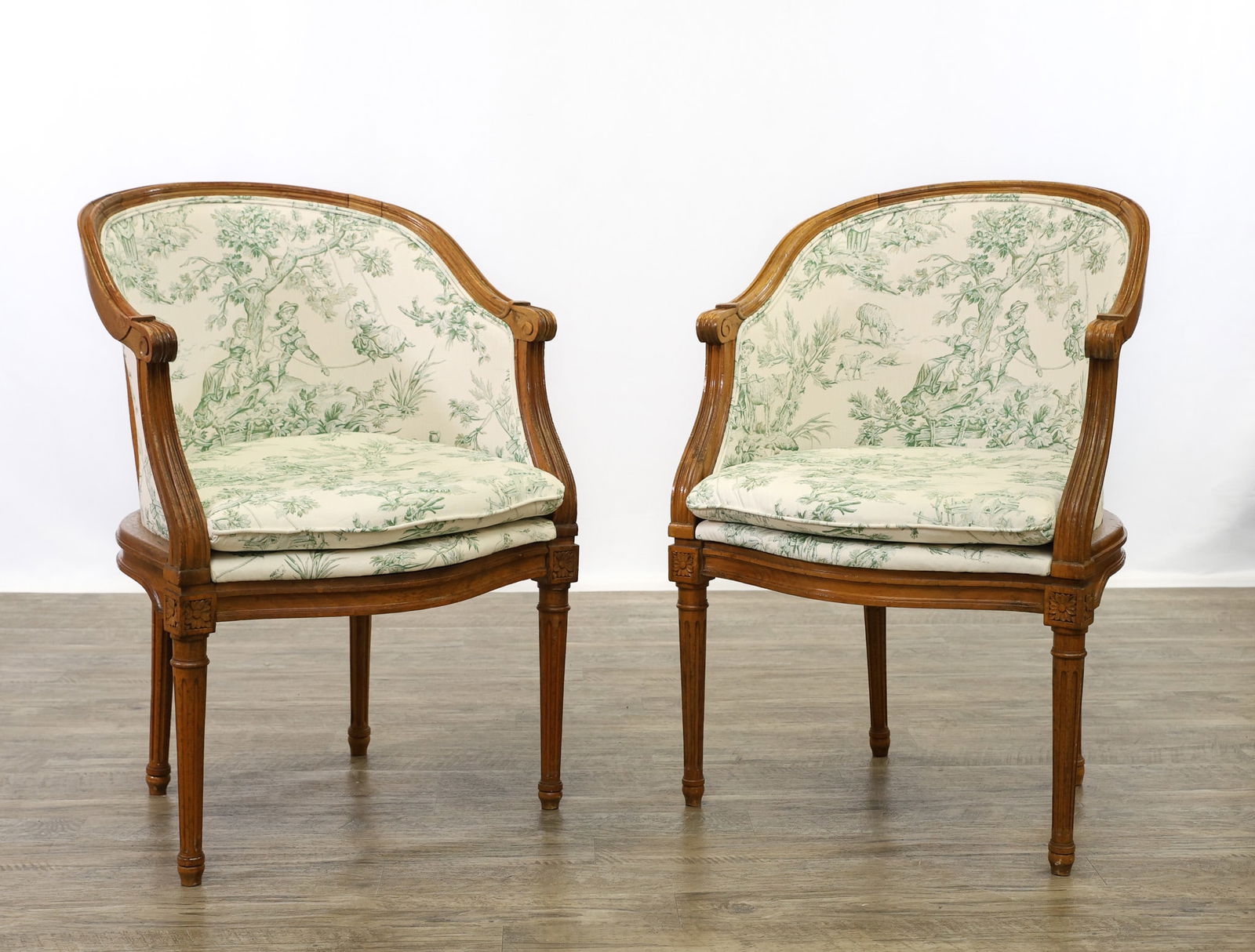 Pair of Louis XVI Accent Chairs: Pair of bergère style chairs with exposed wood frame, rounded back and continuous arm. Exposed mahogany frames and upholstered seat and back. Tapered and fluted legs, front with flower carving detail