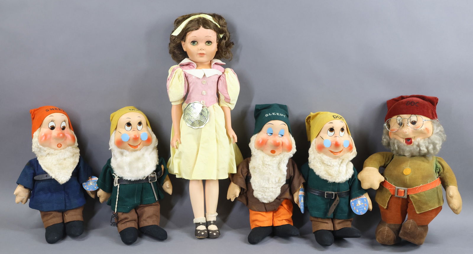 CLOTH DOLLS & SNOW WHITE.: LOT. 21" Tagged "Snow White" - 1950's Vinyl doll by DeLuxe Toy Creations, NY. All original. Condition: age discoloration. (5) Dwarfs - (3) Tagged 12" Ideal Novelty & Toy Co. "Doc", "Sneezy", "Sleepy".