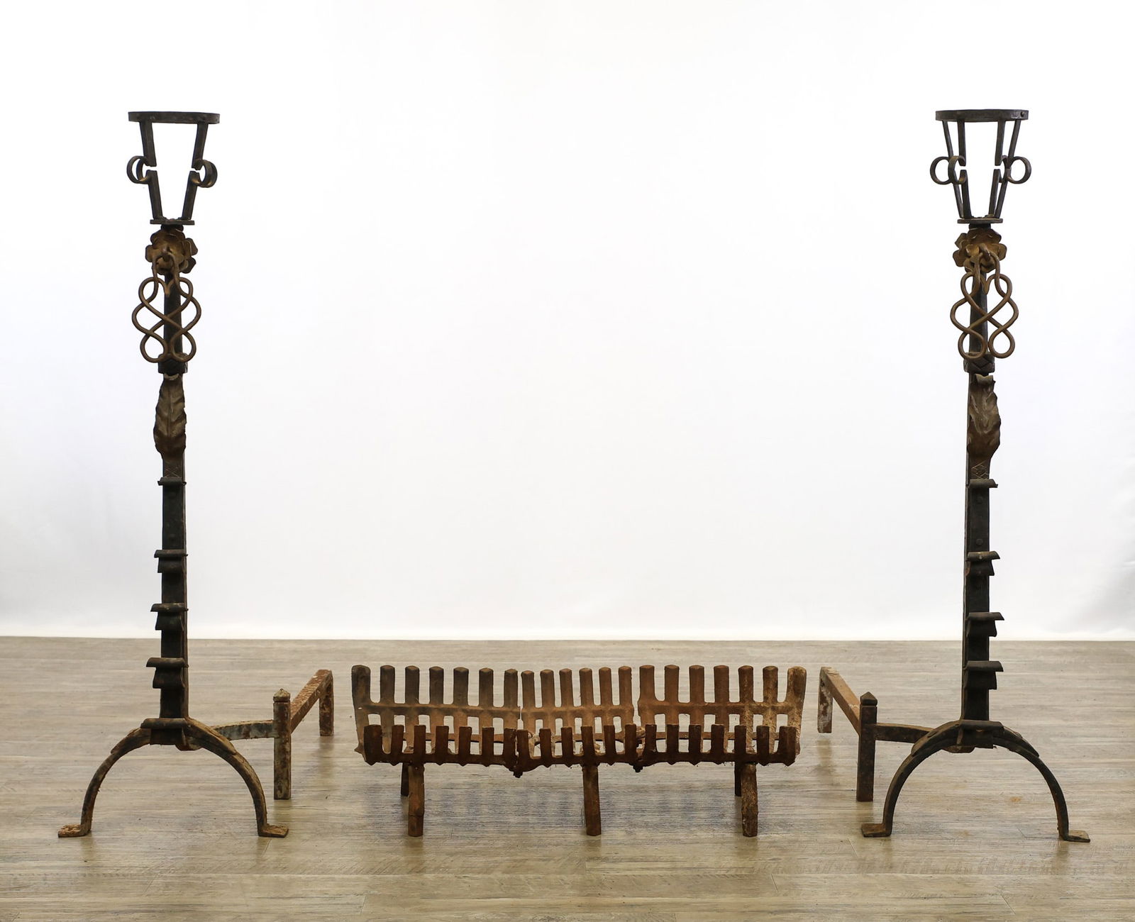 Wrought Iron Andirons in the Manner of Samuel Yellin: Early 20th c. hand-forged with torchiere style tops, floral and acanthus decoration, arched legs with flat feet. 45"h x 14"w x 34"d. Complementing log grate 8"h x 32"w x 15"d