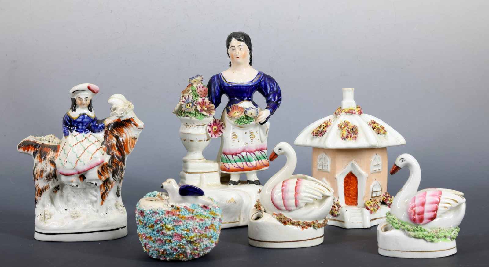 Staffordshire Figures and Pastille Burner: Figure of a woman with floral basket and vase, a figure of a girl on a large dog, a pair of swan spill vases, a small cottage pastille burner, and a colorful encrusted nest spill vase with birds. Larg