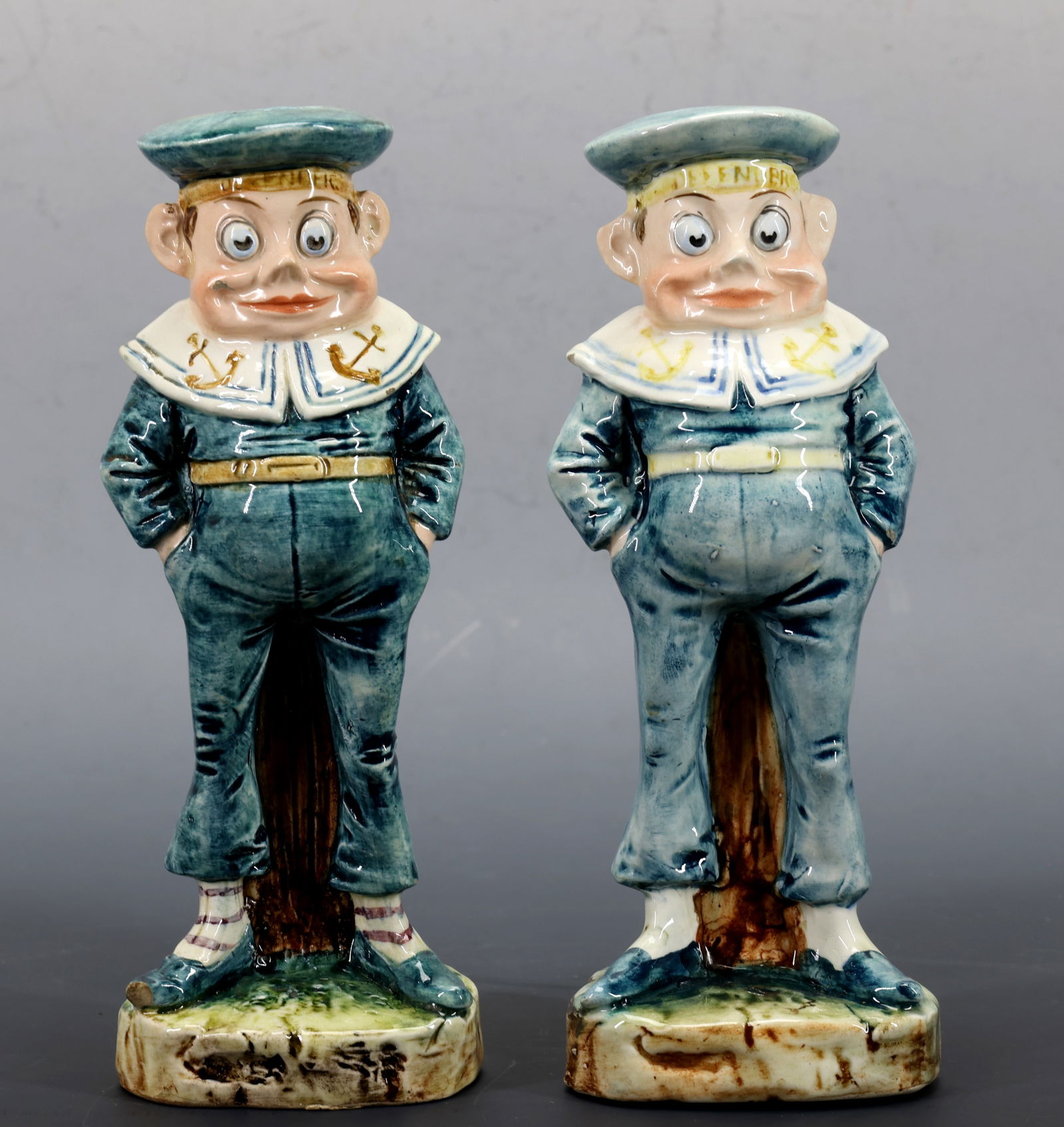 Majolica Figure of The Sailor Defender Brownie from Palmer Cox: Majolica figurines of a Brownie as Sailor Defender standing upon a base of stony ground; wearing a blue sailor uniform and cap which is labeled "Defender", and his cap has an open top designed for use